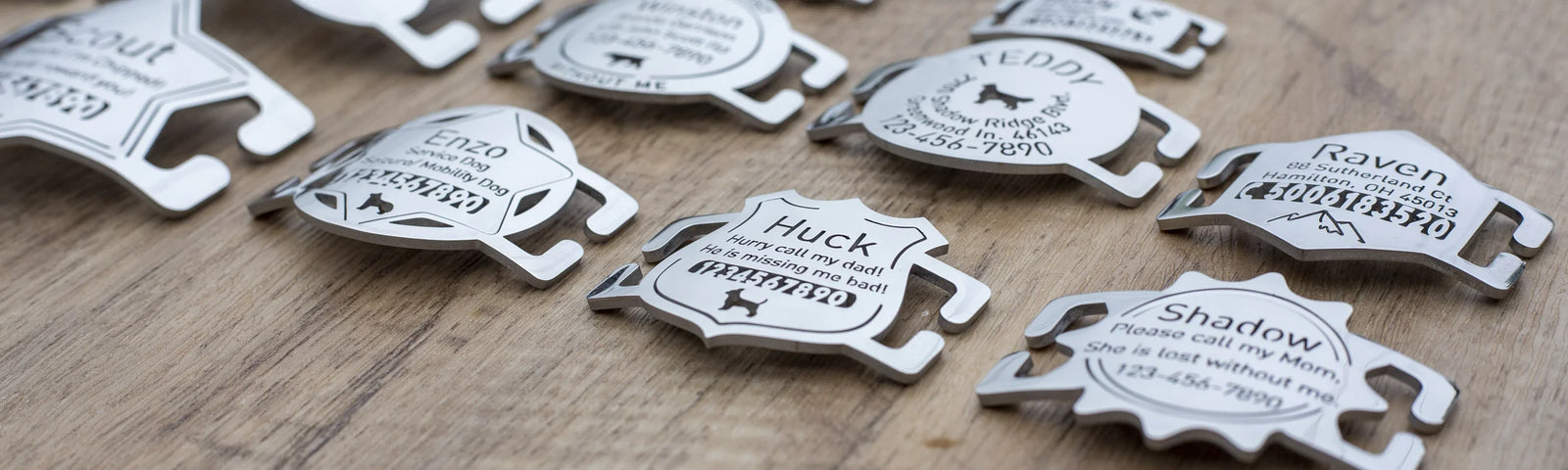 Several small silver metal tags or charms with engraved names and dog illustrations are arranged on a wooden surface.